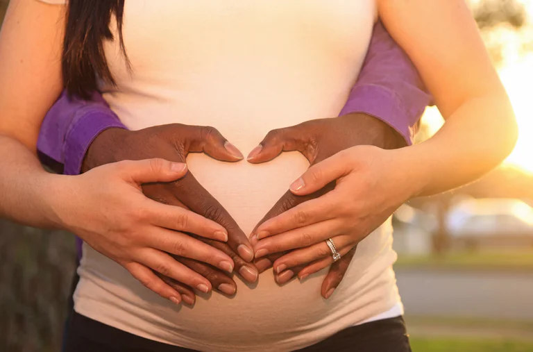 Händer som formar ett hjärta över en gravid mage utomhus i solljus.