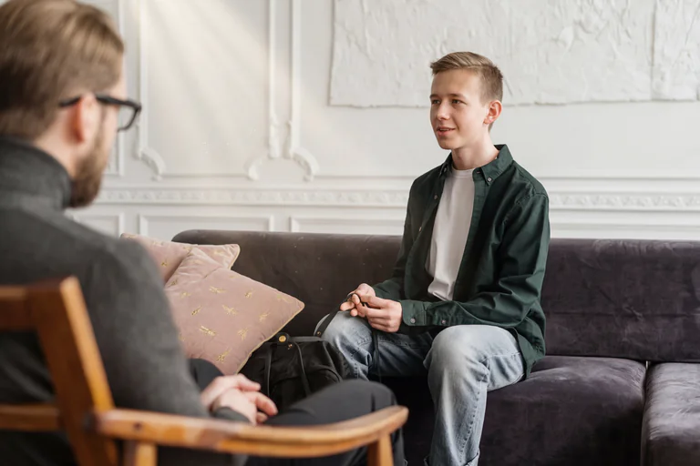 En manlig terapeut med glasögon sitter i en stol och talar med en tonårspojke som sitter i soffan i ett vardagsrum.