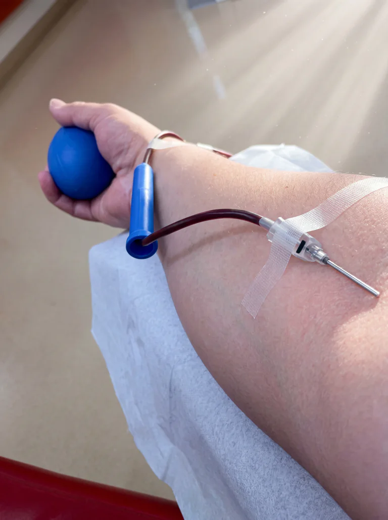 En person som donerar blod, håller en blå stressboll i handen när nålen är instucken i armen.