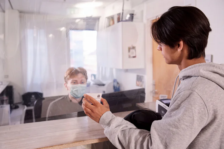 Receptionist bakom en plexiglasskärm tar emot ett ID-kort från en person vid en disk.