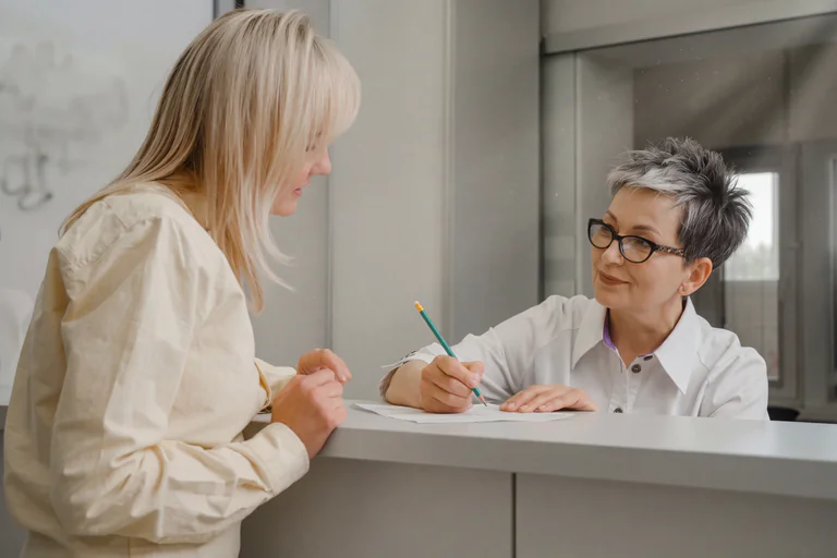 En receptionist med glasögon skriver på ett papper vid ett skrivbord medan en patient står framför och tittar.