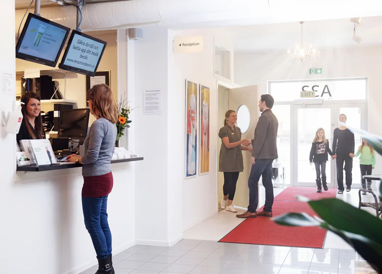 En receptionist bakom en disk hjälper en kund som står framför. I bakgrunden finns en reception och en väntrumsmiljö med personer.