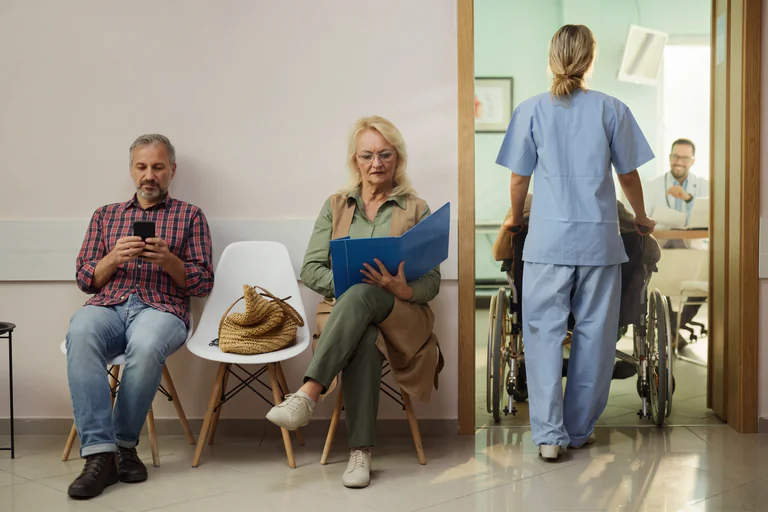 Sjuksköterska skjuter en patient i rullstol genom en korridor i en väntsal, medan två patienter sitter och väntar.