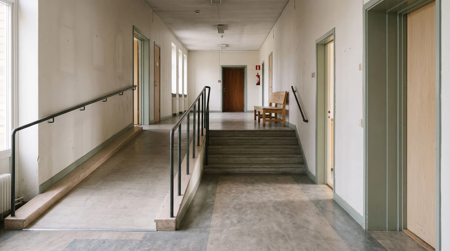 Tom korridor med ljusa väggar. Till vänster leder en ramp med metallräcke uppåt, parallellt med en kort trappa. Längre fram i korridoren står en träbänk intill en stängd dörr.