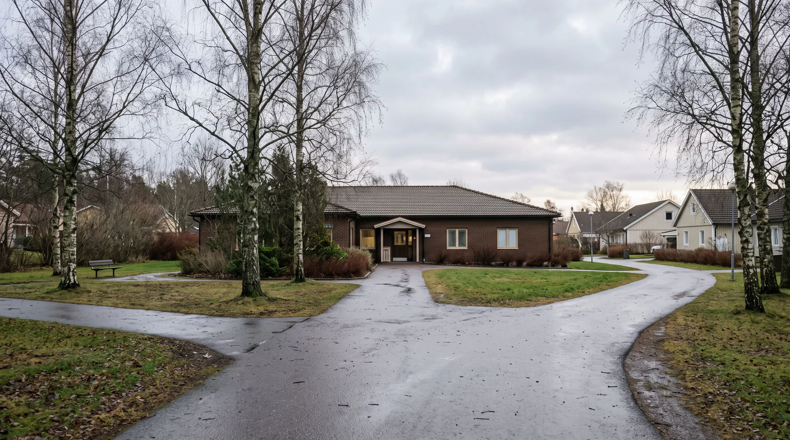 En asfalterad gångväg delar sig i ett vägskäl framför en låg tegelbyggnad omgiven av kala björkar och gräsmattor.
