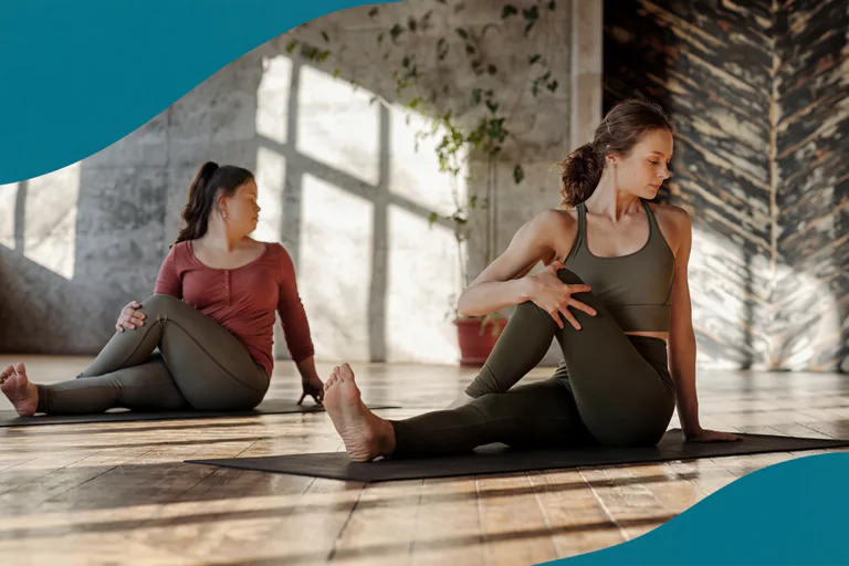 Två kvinnor utför en vridningsposition i yoga under ett soligt pass i en studio.