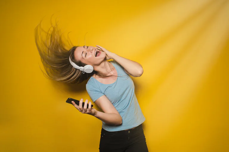 En kvinna med hörlurar på huvudet och en mobiltelefon i handen dansar glatt med håret i luften mot en gul bakgrund.