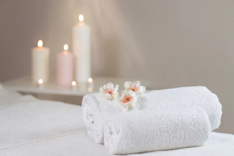 Vita hoprullade handdukar med blommor på och tre ljus i bakgrunden i en spa-miljö.
