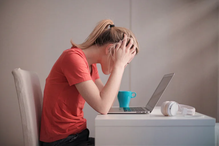 En kvinna med händerna på huvudet sitter framför en laptop och ser frustrerad ut, med en mugg och hörlurar bredvid.