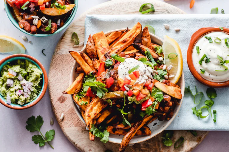 En skål med sötpotatispommes frites toppad med guacamole, salsa, gräddfil och örter, serverad med en skål guacamole och en skål gräddfil på ett träunderlag.