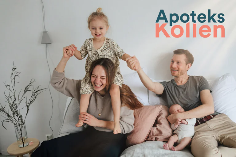 En leende familj med två barn sitter på en soffa hemma, med en baby i famnen och en flicka som sitter på mammas axlar.