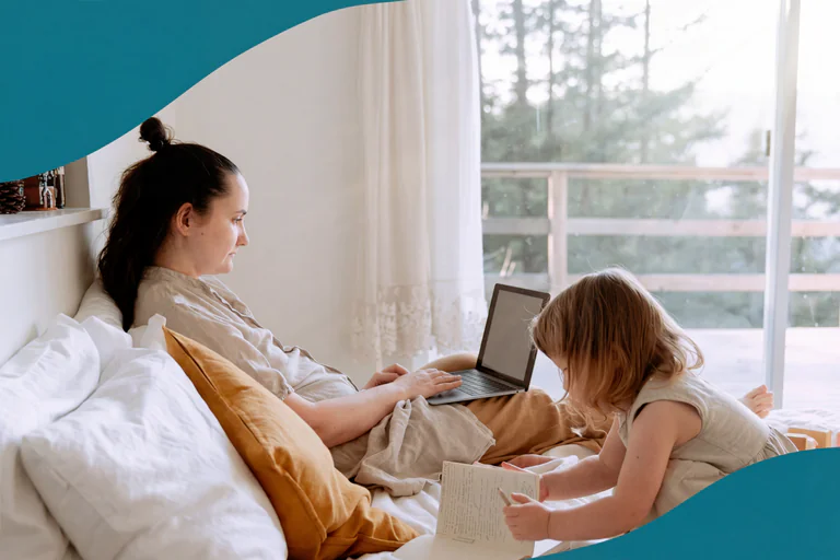 En mamma sitter på en säng med en laptop och arbetar medan hennes lilla barn sitter bredvid och läser en bok.