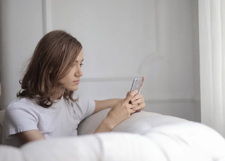 En kvinna sitter på en soffa och tittar på sin smartphone, med en gardin i bakgrunden.