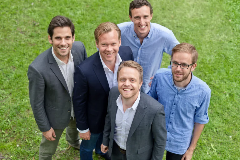 En grupp av fem män står utomhus på gräs och ler mot kameran.