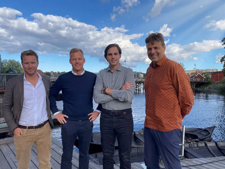 En grupp på fyra män står tillsammans utomhus vid en vattensamling med en bro och byggnader i bakgrunden.