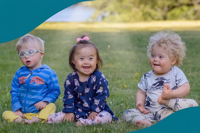 Tre barn sitter på gräset utomhus, två med Downs syndrom, och ler.