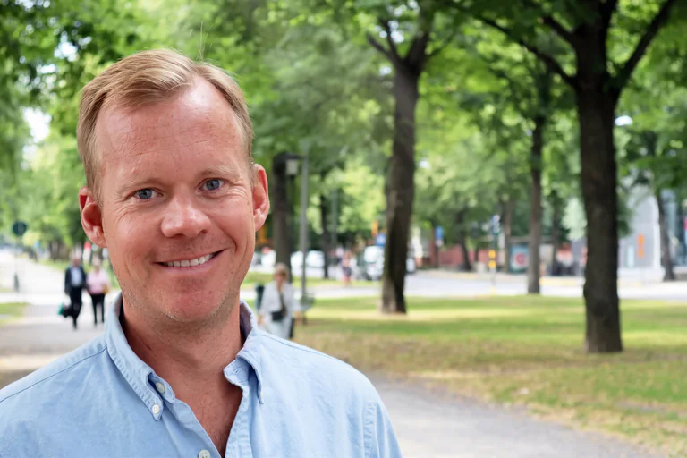 En leende man i ljusblå skjorta står utomhus i en park med träd och människor i bakgrunden.