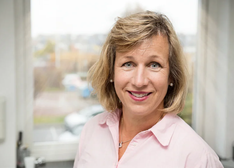 Porträtt av en medelålders kvinna med blont hår som ler och tittar in i kameran, inomhus.