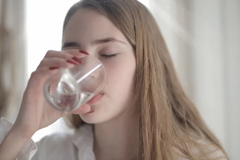En ung kvinna med långt blont hår dricker vatten ur ett glas.