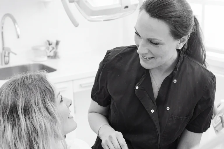 Tandläkare i mörk klädsel som ler och pratar med en patient med ljusblont hår i en klinik.