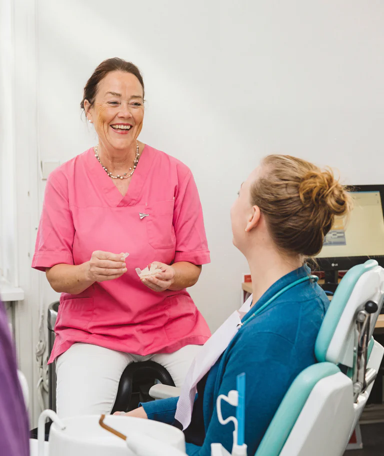 Tandläkare i rosa arbetskläder förklarar något för en patient som sitter i en tandläkarstol.