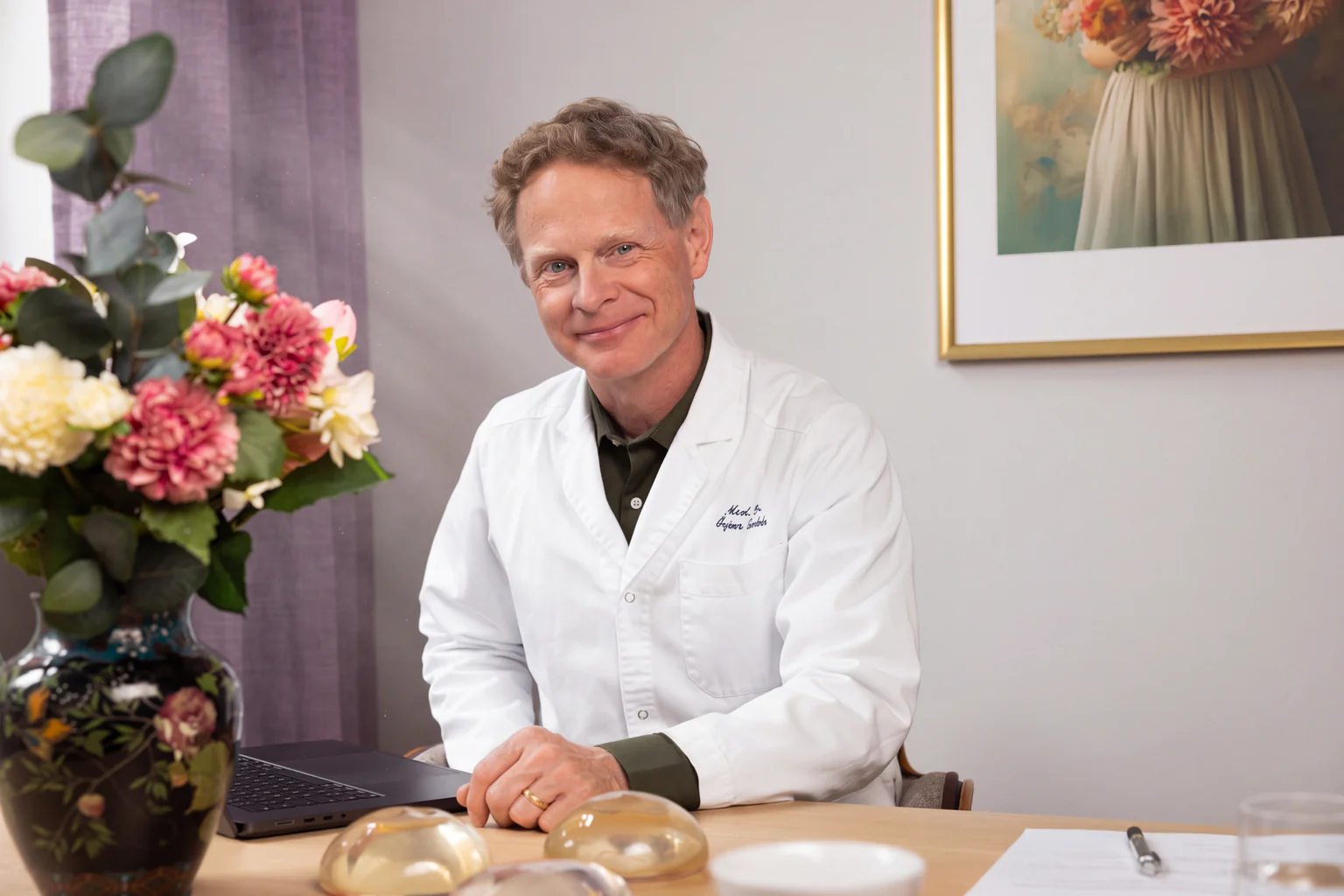 Man i läkarrock sitter vid ett skrivbord med en blomsterbukett och medicinska implantat
