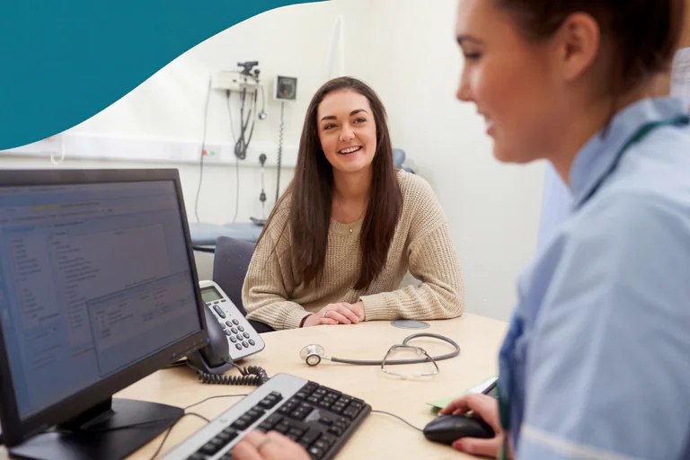 En leende patient sitter vid ett skrivbord och tittar på en läkare som sitter mittemot henne. På skrivbordet finns en datorskärm, ett stetoskop och en telefon.