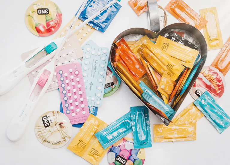 En platt lagg av olika preventivmedel, inklusive kondomer i olika färger och förpackningar, p-pillerkartor och graviditetstest.