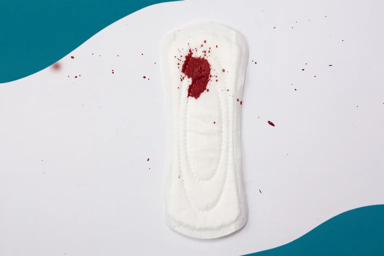 Ett vitt hygieniskt skydd med en blodfläck i mitten och flera små blodstänk runt omkring, placerat på en vit bakgrund med en turkos vågig kant.