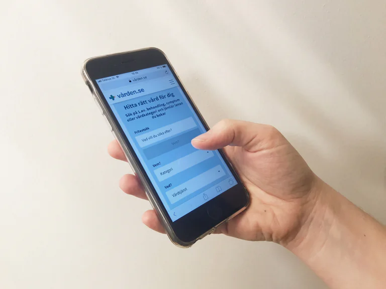 En hand som håller i en smartphone och navigerar på en svensk vårdplattform.